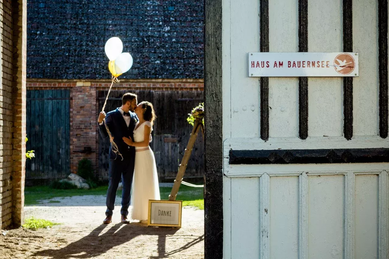 Hochzeitsfotos Haus am Bauernsee:Ein Hochzeitspaar steht sich küssend in einer rustikalen Scheune gegenüber. Der Bräutigam hält bunte Luftballons, während ein Schild mit der Aufschrift "Haus am Bauernsee" im Vordergrund zu sehen ist.