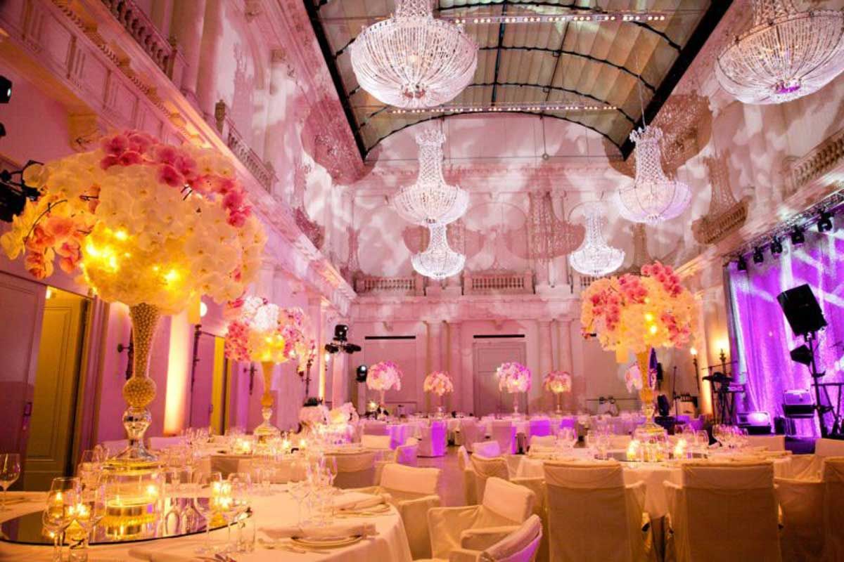 Ein prächtig dekorierter Hochzeitssaal im Hotel de Rome in Berlin mit hohen Decken, großen Kristallleuchtern und festlich gedeckten Tischen mit Blumenarrangements und Kerzen.