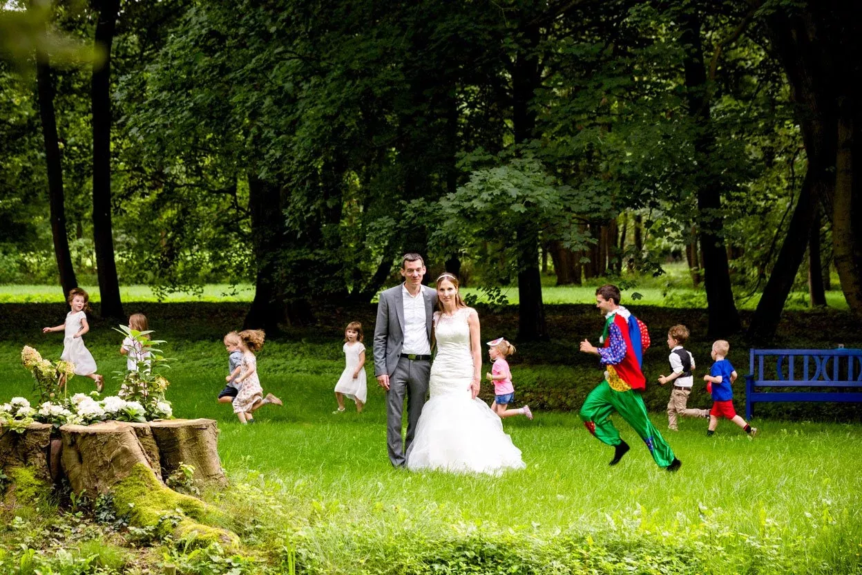 Braut und Bräutigam stehen inmitten einer Gruppe von spielenden Kindern und einem farbenfrohen Unterhalter in einem Waldpark. Die Szene spiegelt Freude und Gelassenheit wider.