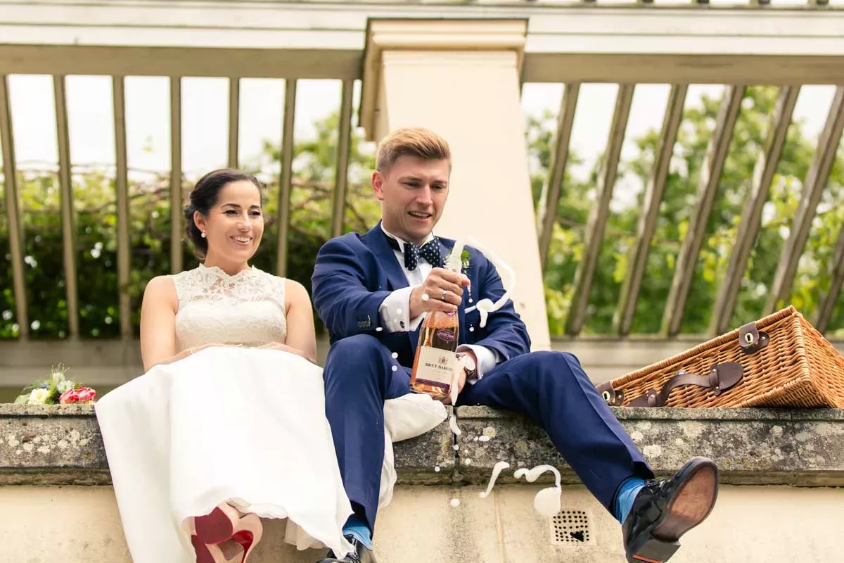 Ein fröhlicher Moment während einer Hochzeit im Schloss Glienicke: Das Brautpaar öffnet Champagner unter einer Pergola.