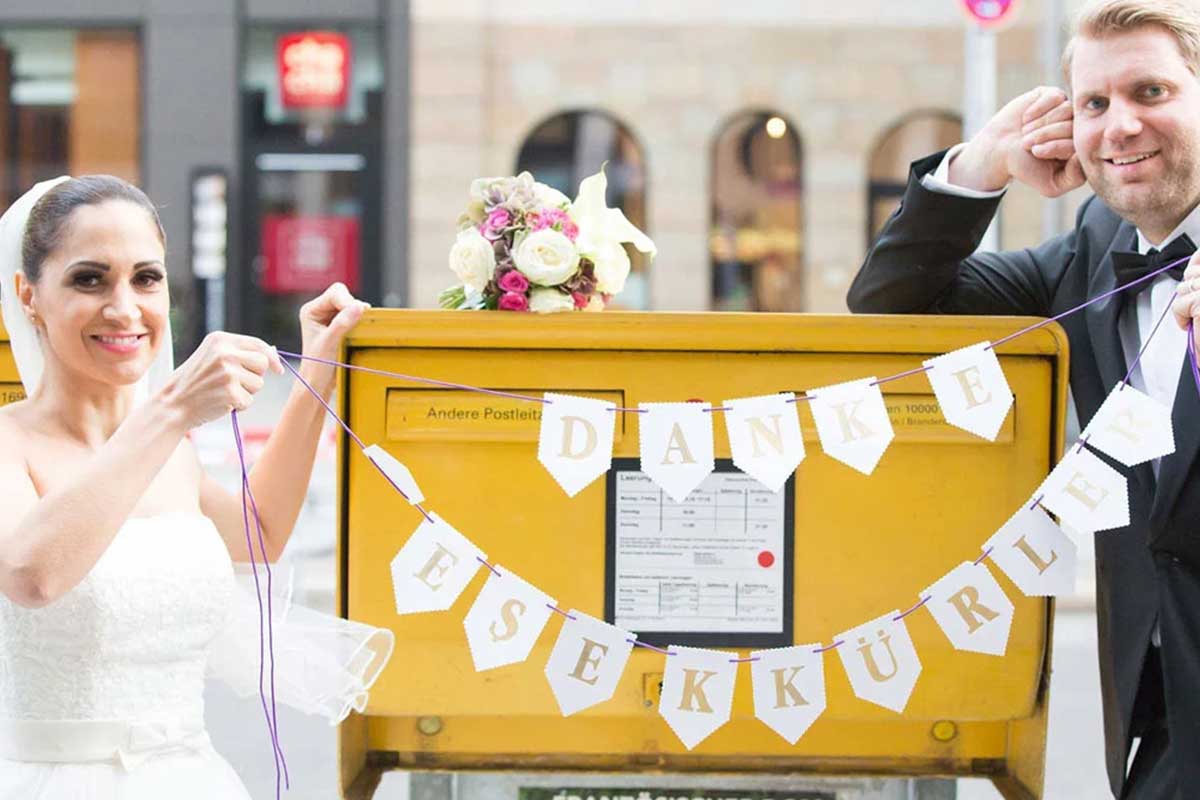 Brautpaar hält ein Banner mit der Aufschrift "DANKE" und "TEŞEKKÜRLER" vor einem gelben Postkasten.