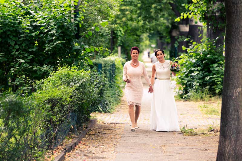 Eine Braut und ihre Mutter, gekleidet in ein weißes Hochzeitskleid und ein cremefarbenes Spitzenkleid, gehen zusammen auf einem von Bäumen umsäumten Weg zum Standesamt Schmargendorf, umgeben von einer ruhigen, grünen Landschaft.
