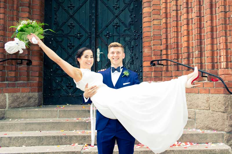 Die Braut schwingt ihr Hochzeitskleid hoch, während sie vom Bräutigam vor dem Standesamt Schmargendorf gehalten wird, beide strahlen vor Glück.