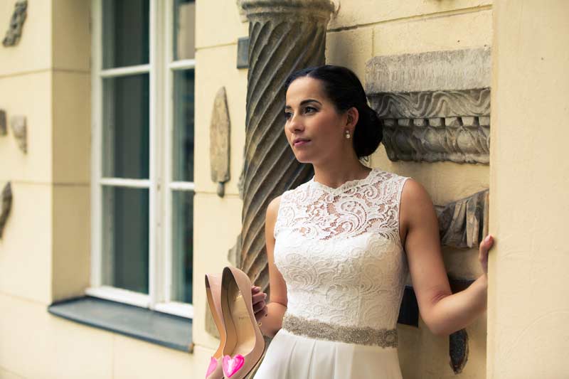 Braut in einem weißen Spitzenkleid steht neben einem historischen Fenster bei Schloss Glienicke und hält ihre Schuhe, während sie in die Ferne blickt.