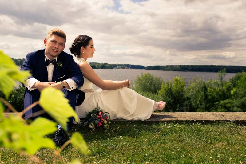  Frisch vermähltes Paar sitzt am Ufer eines Sees nahe Schloss Glienicke, umgeben von grüner Natur und mit Blick auf das Wasser, Bräutigam in blauem Anzug und Braut in weißem Kleid.
