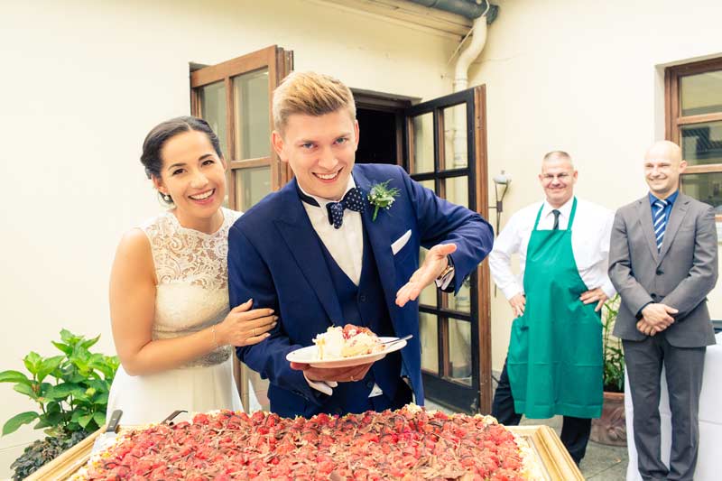 Das Brautpaar im Schloss Glienicke lacht, während der Bräutigam seiner Braut ein Stück Erdbeertorte anbietet, umgeben von einem großen Erdbeerkuchen und beobachtet vom lächelnden Servicepersonal.
