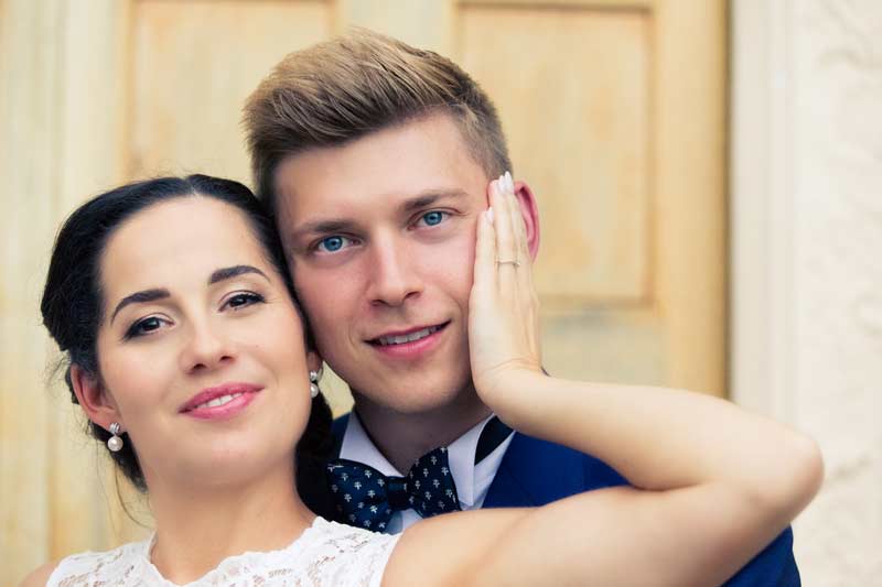 Die Braut, gekleidet in einem weißen Spitzenkleid, hält liebevoll das Gesicht des Bräutigams in einem blauen Anzug, beide lächeln glücklich.