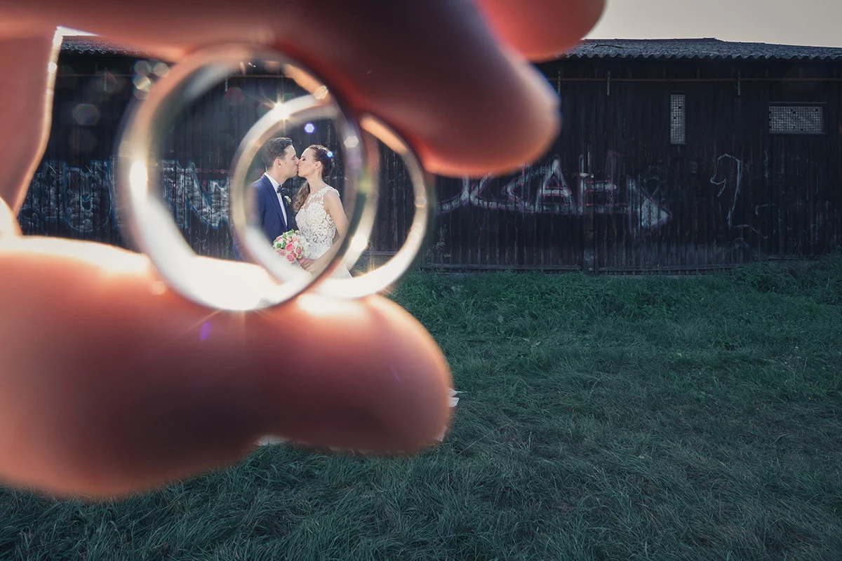 Ein Brautpaar, das durch die Linse eines Verlobungsrings betrachtet wird, steht vor einer rustikalen Holzwand mit Graffiti und küsst sich.