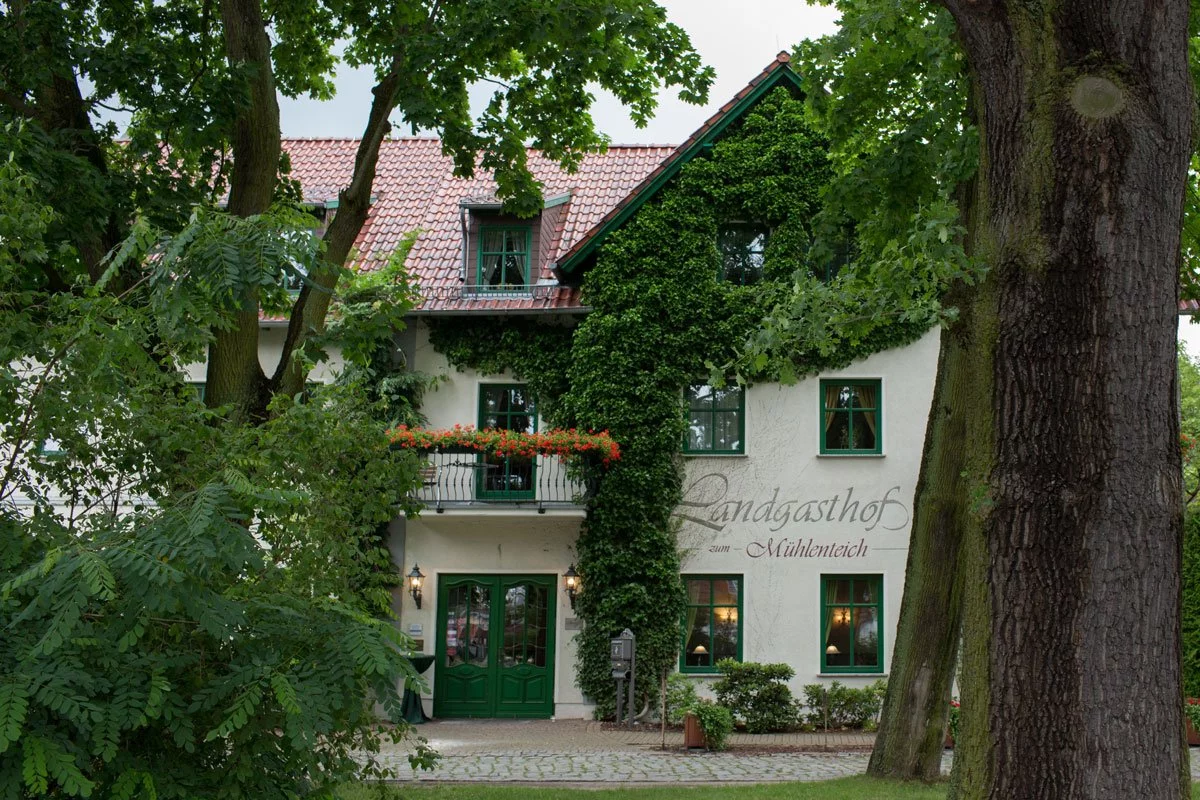 hochzeit Landgasthof zum Mühlenteich: Außenansicht des Landgasthofs zum Mühlenteich, umgeben von üppigem Grün und alten Bäumen, mit einem charmanten, bewachsenen Äußeren und roten Dachziegeln.