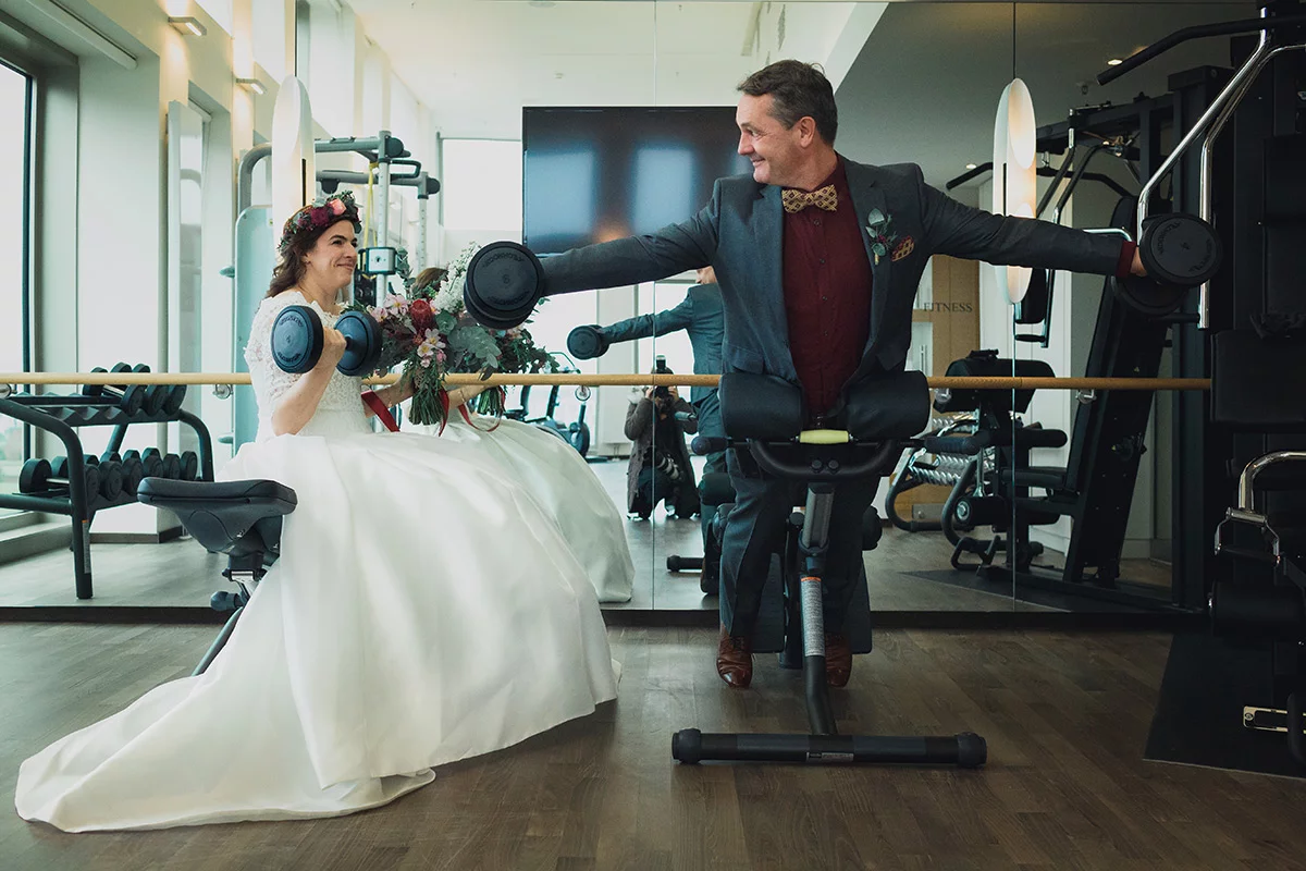 Braut und Bräutigam im Fitnessstudio der Villa Blumenfisch, die Hanteln heben und lachen.