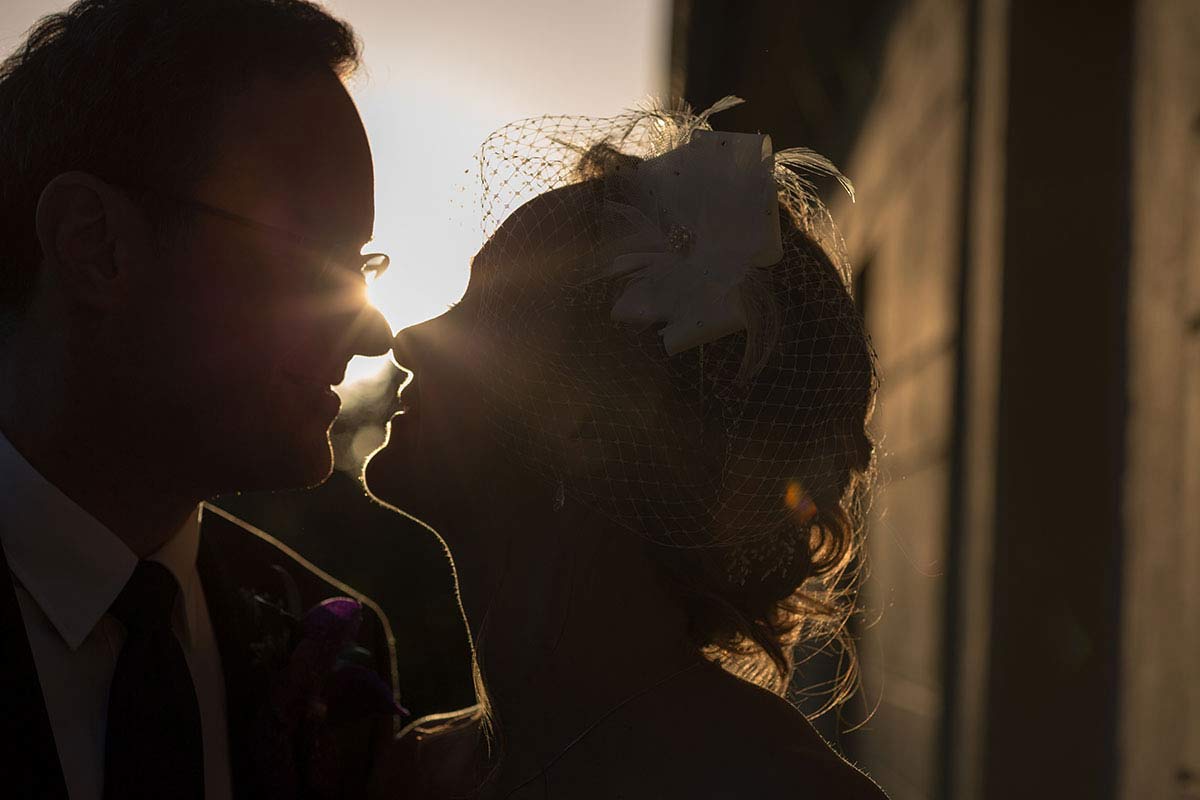 Das Brautpaar während eines romantischen Paar-Shootings bei Sonnenuntergang. Die Silhouetten sind im Gegenlicht der untergehenden Sonne zu sehen. Die Braut trägt einen Haarschmuck mit Schleier, der Bräutigam einen dunklen Anzug. Die Szene ist stimmungsvoll und romantisch.