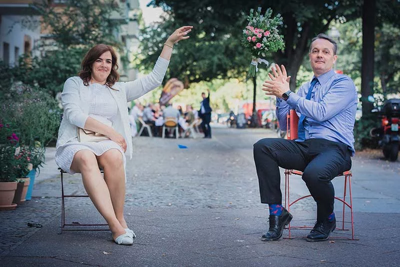 Braut und Bräutigam sitzen auf Stühlen auf einer gepflasterten Straße, wobei die Braut den Arm hebt und der Bräutigam den Blumenstrauß fängt.