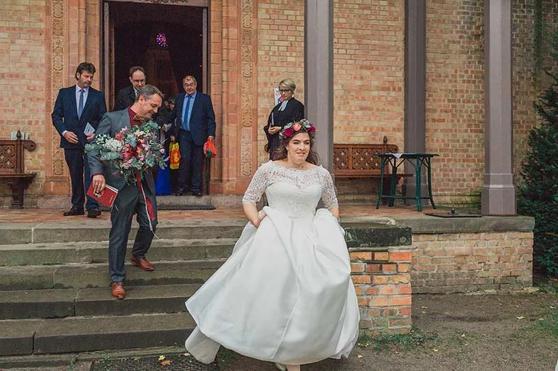 Braut und Bräutigam verlassen die Kirche nach ihrer Hochzeitszeremonie in der Villa Blumenfisch, umgeben von Gästen und der Traurednerin.