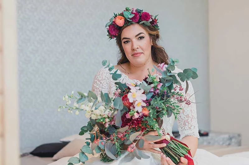 Braut in einem weißen Spitzenkleid mit einem Blumenkranz im Haar hält einen bunten Brautstrauß in der Villa Blumenfisch.