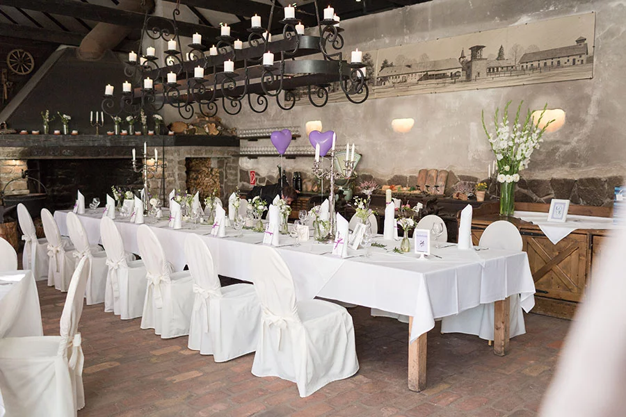Rustikale Hochzeitsdekoration in einem ländlichen Saal im Schloss Diedersdorf, mit weiß gedeckten Tischen, Blumenarrangements und lila Herzballons.