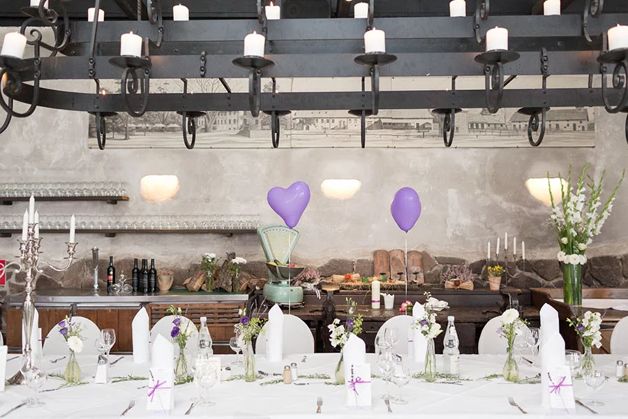 Rustikale Hochzeitsdekoration in einem ländlichen Saal im Schloss Diedersdorf, mit weiß gedeckten Tischen, Blumenarrangements und lila Herzballons.
