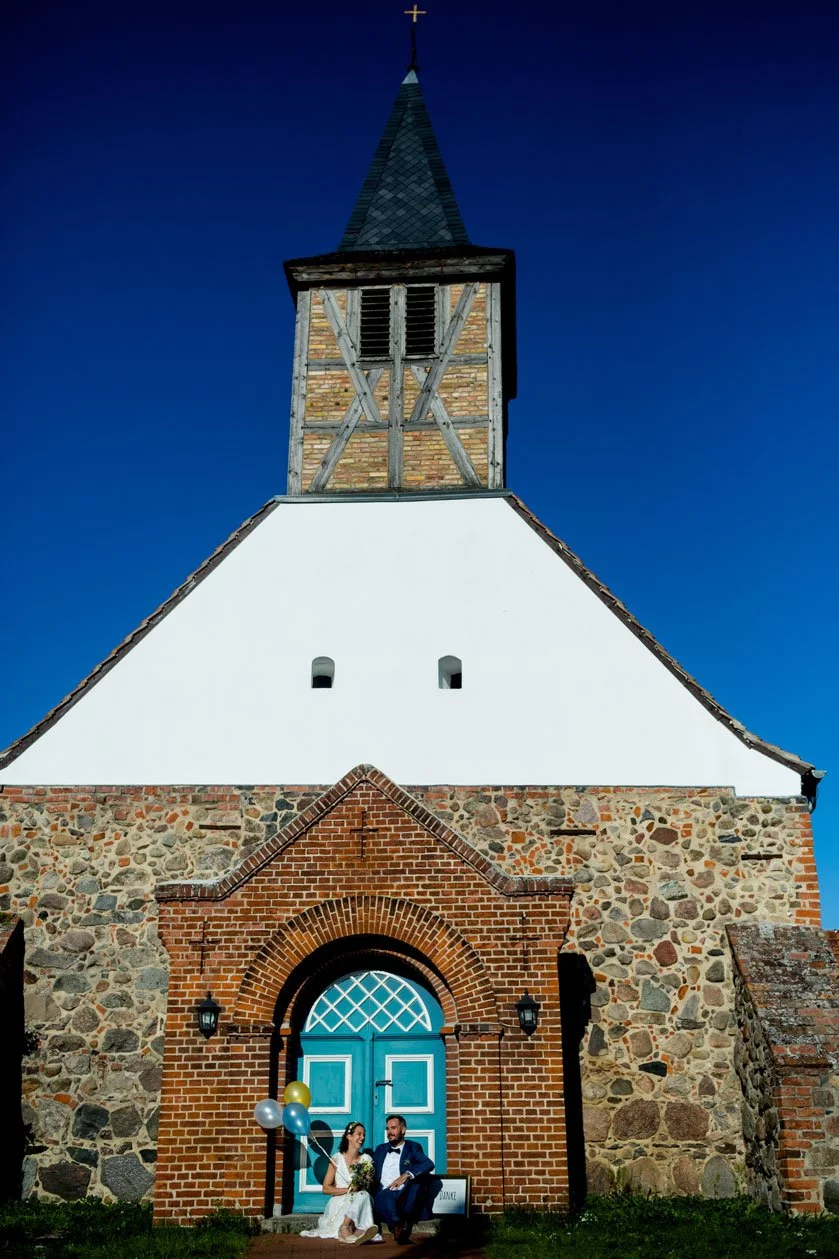 Brautpaar sitzt vor der rustikalen Backsteinkirche mit blauem Eingangstor und festlichen Ballons, unter einem klaren, blauen Himmel.