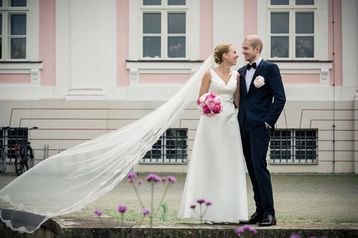  Ein Hochzeitspaar steht lächelnd vor dem Schloss Friedrichsfelde, die Braut in einem weißen Kleid mit Schleier, der Bräutigam in einem blauen Anzug.