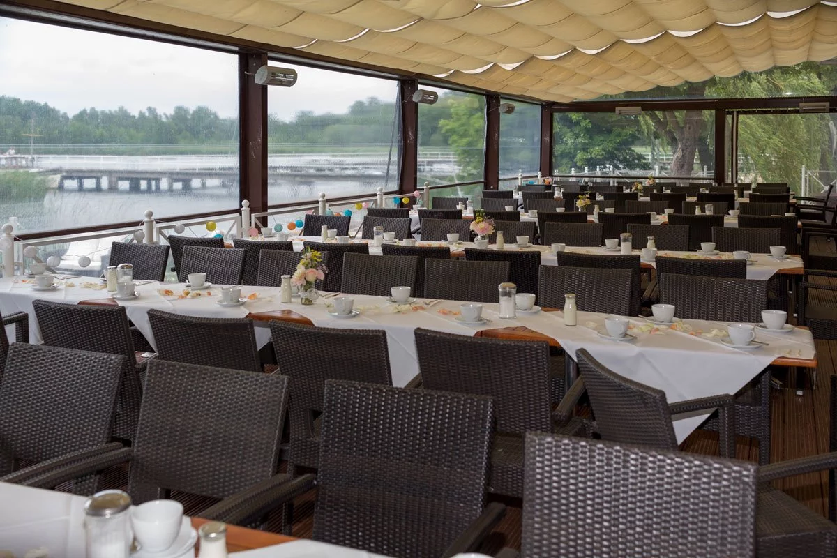 Überdachte Terrasse für eine Hochzeit in der Seelodge Kremmen mit Rattanstühlen, weißen Tischdecken und einem Ausblick auf den See durch breite Fenster.