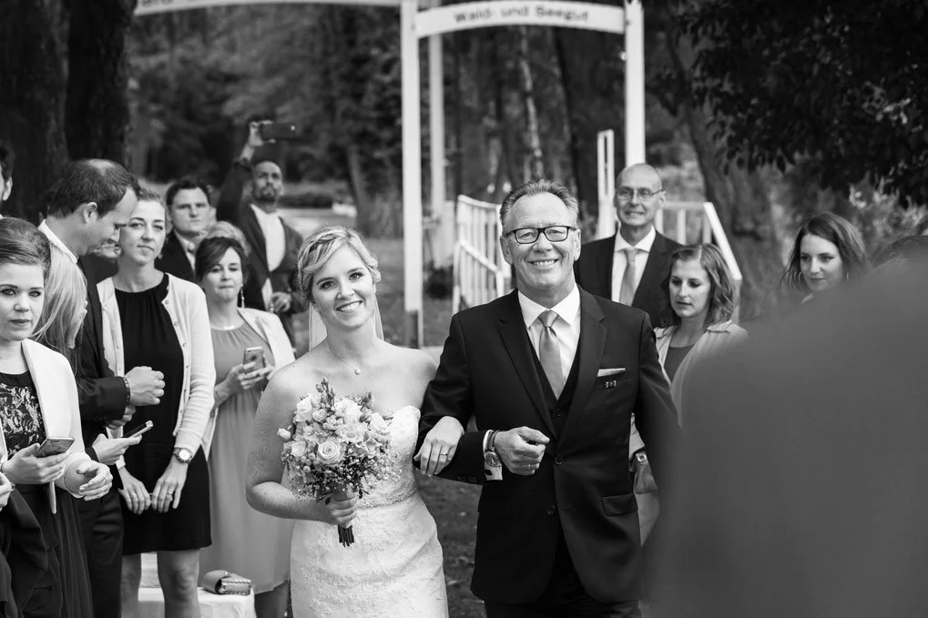 Vater begleitet die Braut zur Hochzeit an der Seelodge Kremmen, umgeben von lächelnden Gästen.