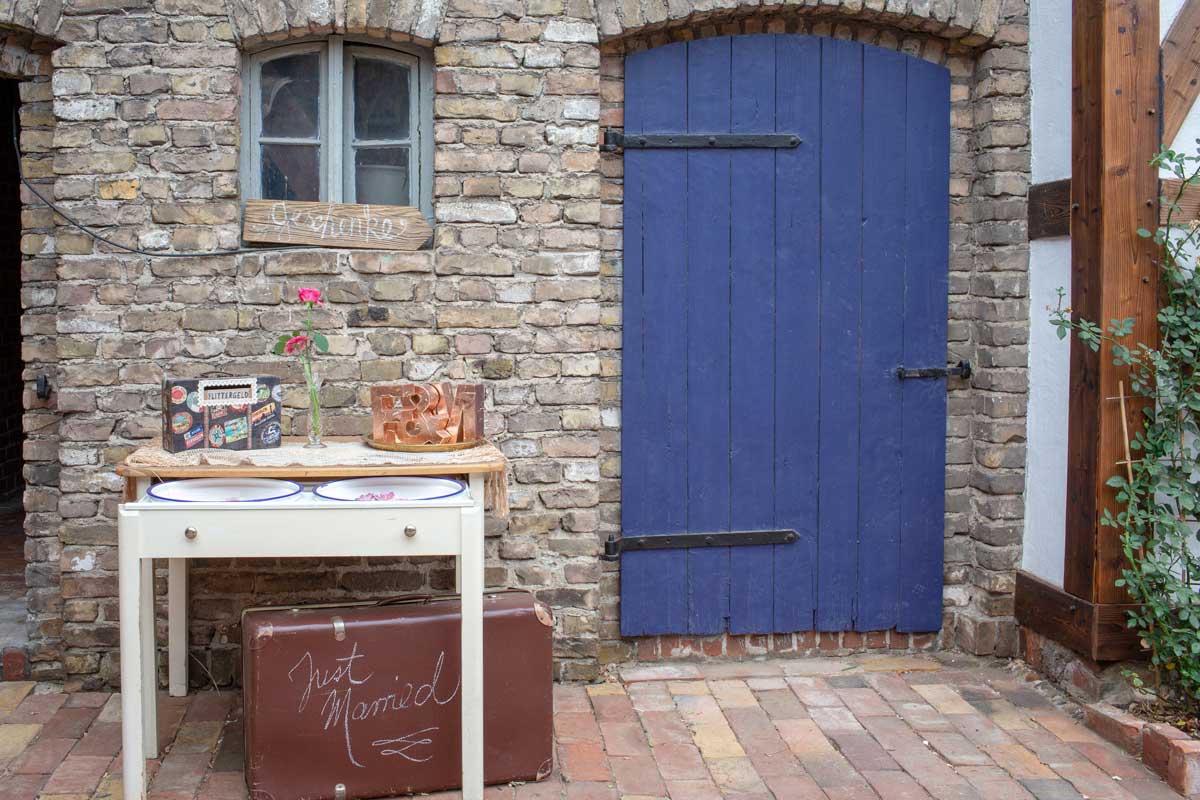 Dekorativer Hochzeitstisch vor einer blauen Holztür in der Hochzeitsscheune Glücksburg, geschmückt mit einer Vase und einer Box für Flittergeld.