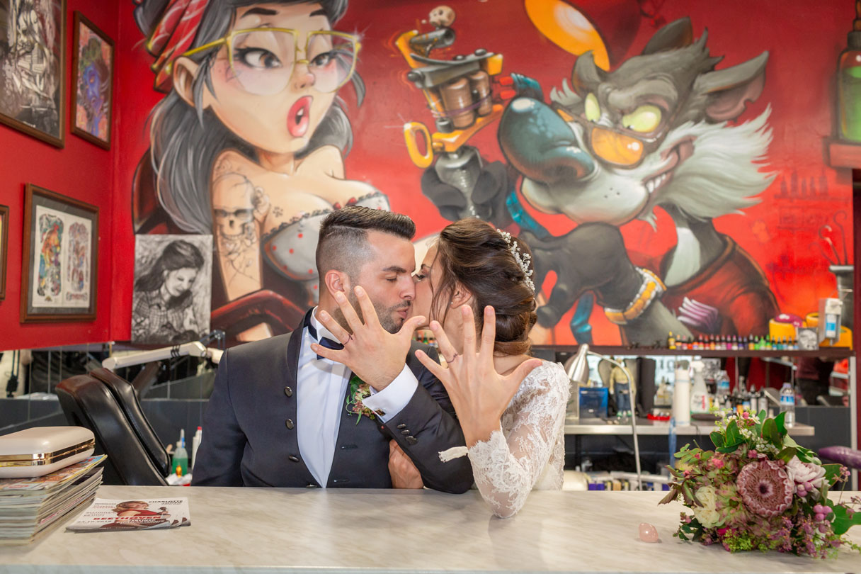 Frisch verheiratetes Paar im Tattoostudio zeigt tätowierte Eheringe vor einer bunten Graffiti-Wand.