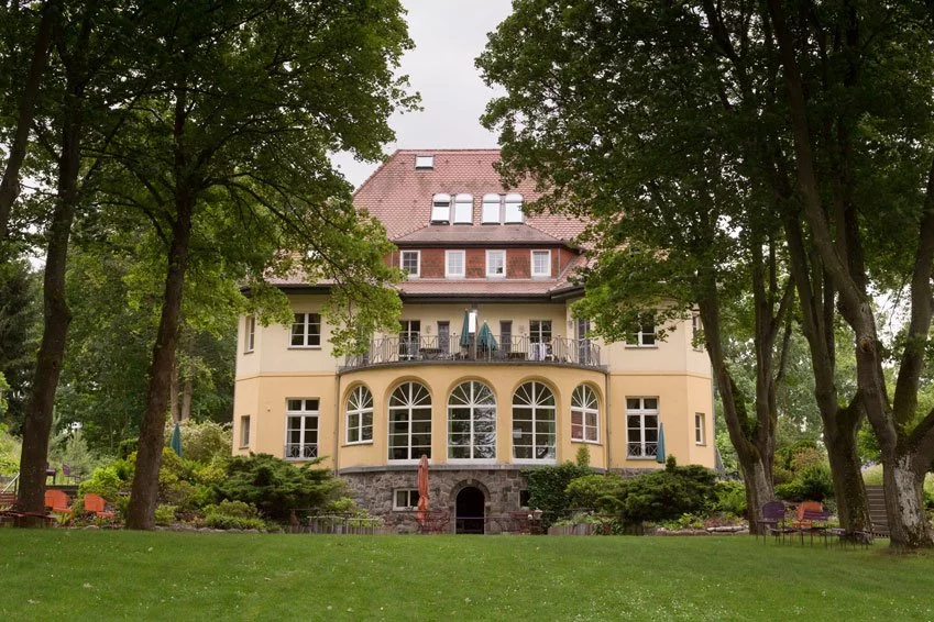 Das Landhaus Himmelpfort am See, umgeben von Bäumen und einem gepflegten Garten, mit einem großen Balkon und historischen Fenstern.