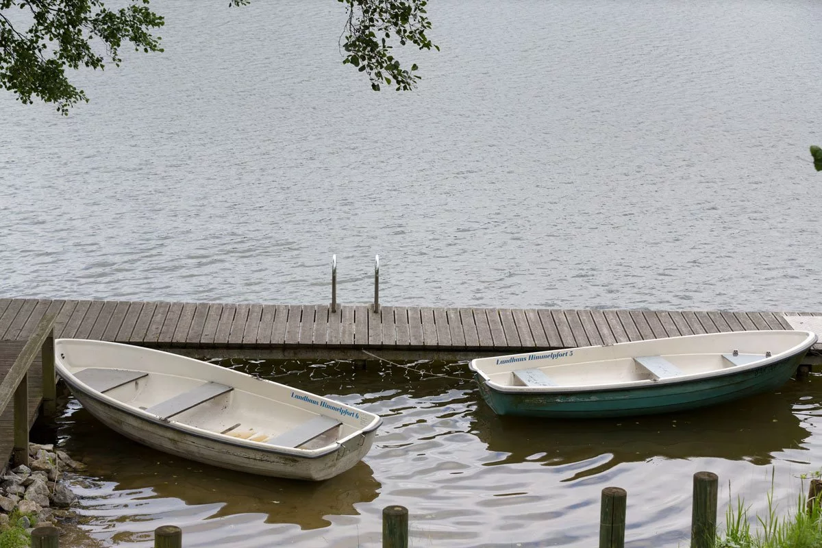 Zwei leere Ruderboote sind an einem hölzernen Steg im See des Landhauses Himmelpfort festgemacht, umgeben von ruhiger Natur.