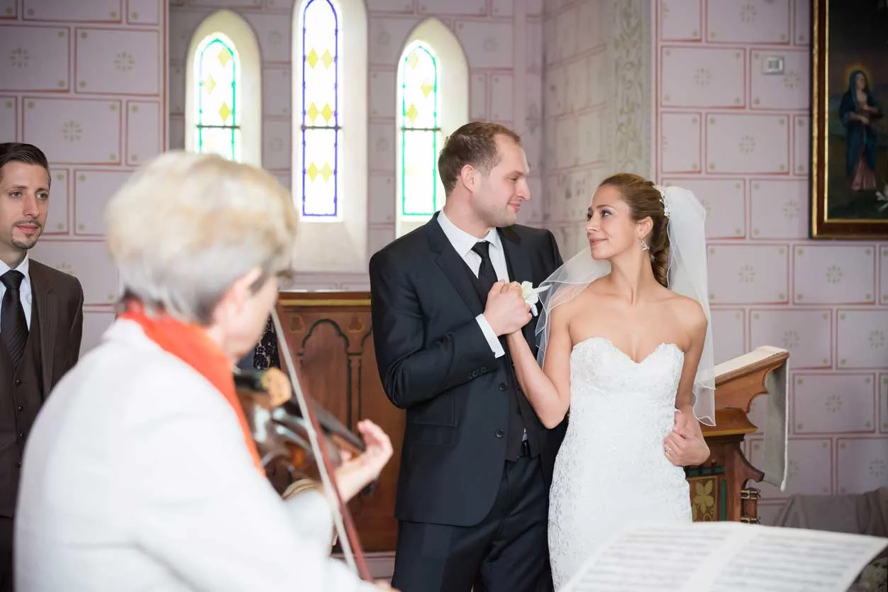 Das Brautpaar teilt einen emotionalen Blickwechsel während der Hochzeitszeremonie, mit einer Violinistin im Vordergrund und bunten Kirchenfenstern im Hintergrund, Hochzeitsfotos Schloss Kartzow.