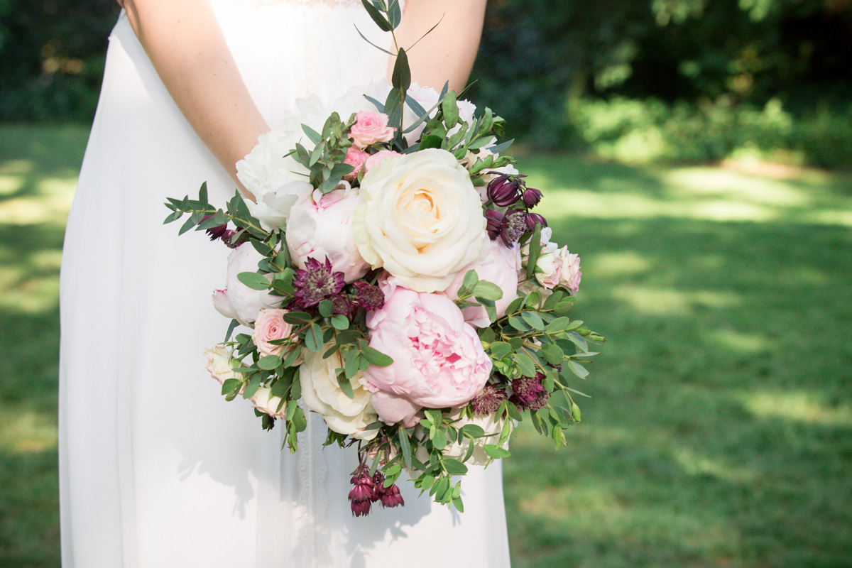 Nahaufnahme des Brautstraußes, den die Braut liebevoll in den Händen hält, mit weißen, rosa und lila Blumen und üppigem Grün.

