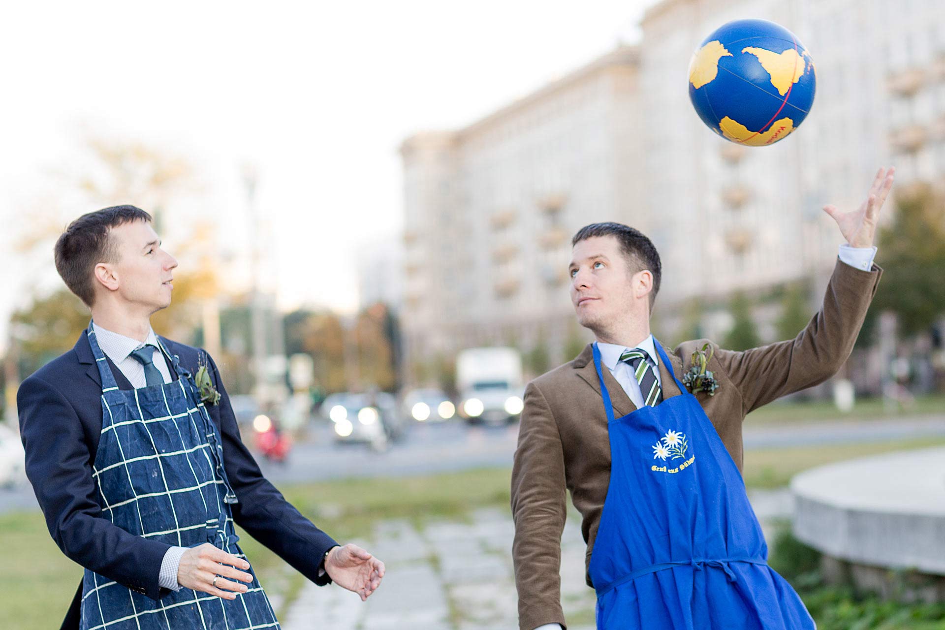 Brautpaar steht auf einem städtischen Platz, trägt bunte Schürzen und spielt mit einem Globusball.
