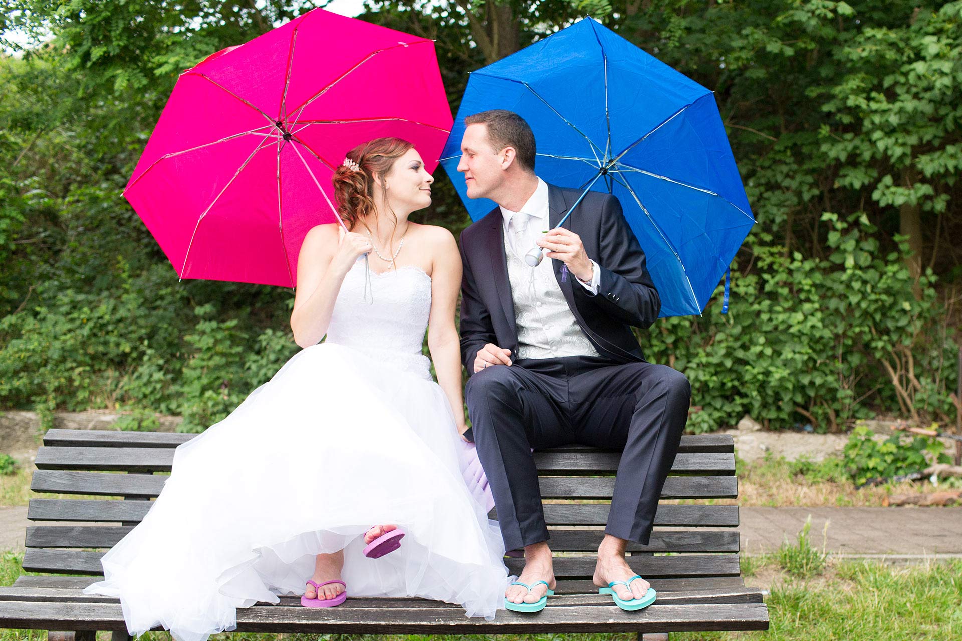 Brautpaar sitzt auf einer Bank und hält pinken und blauen Regenschirm, beide tragen passende Flip-Flops.