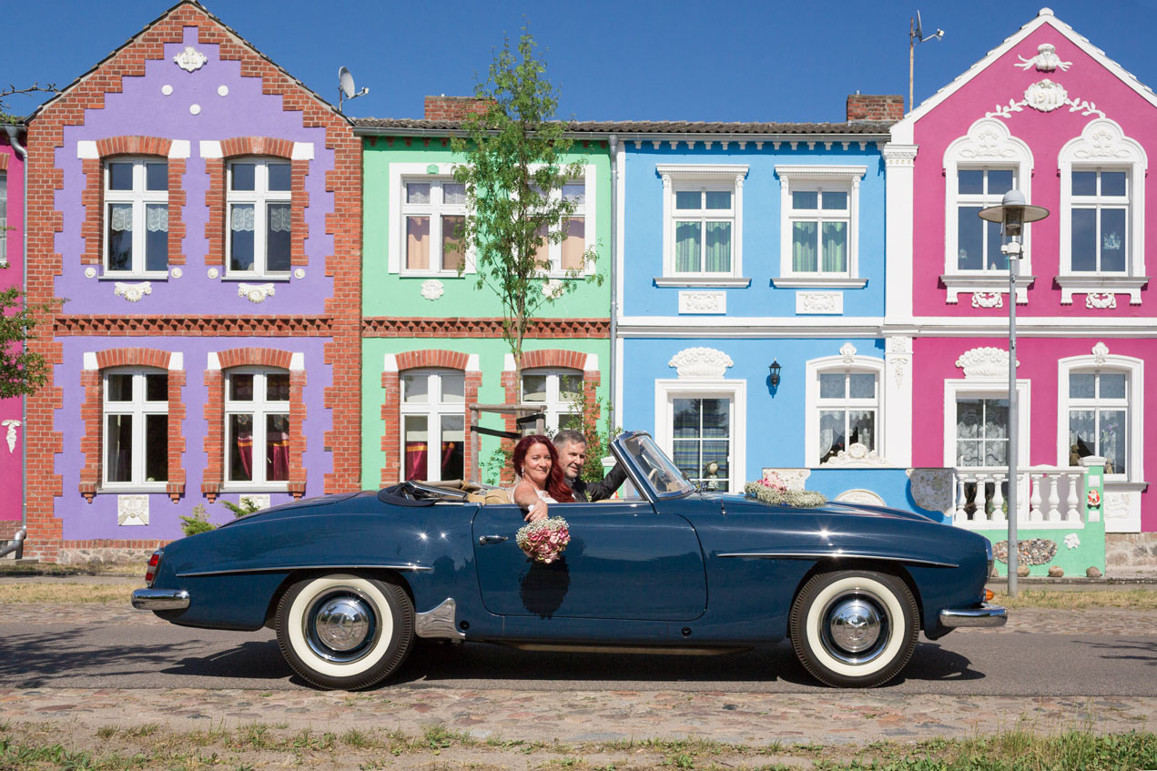 Braut und Bräutigam im Oldtimer-Cabrio vor farbenfrohen Häusern.